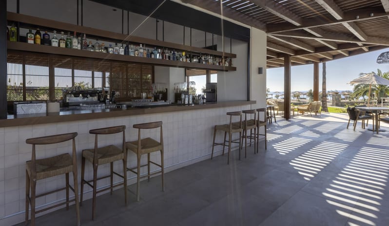 The Spiritzeria tiled counter, woven wooden bar stools, and a wooden slat roof casting shadows over a bright terraceat Aulūs Lindos Rhodes, Curio Collection by Hilton by Kenwood Travel