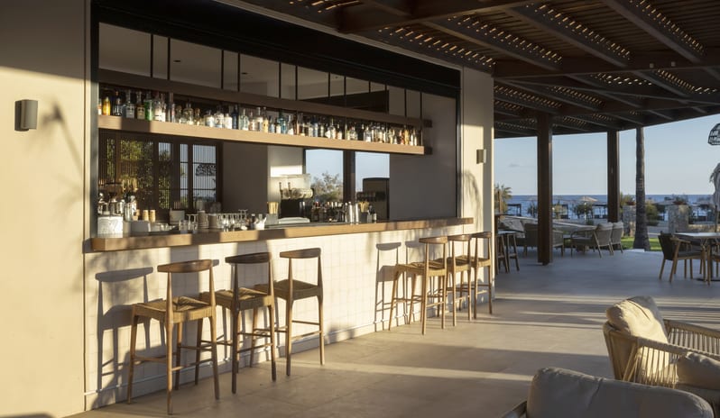 The Spiritzeria tiled counter, woven wooden bar stools, and a wooden slat roof casting shadows over a bright terraceat Aulūs Lindos Rhodes, Curio Collection by Hilton by Kenwood Travel