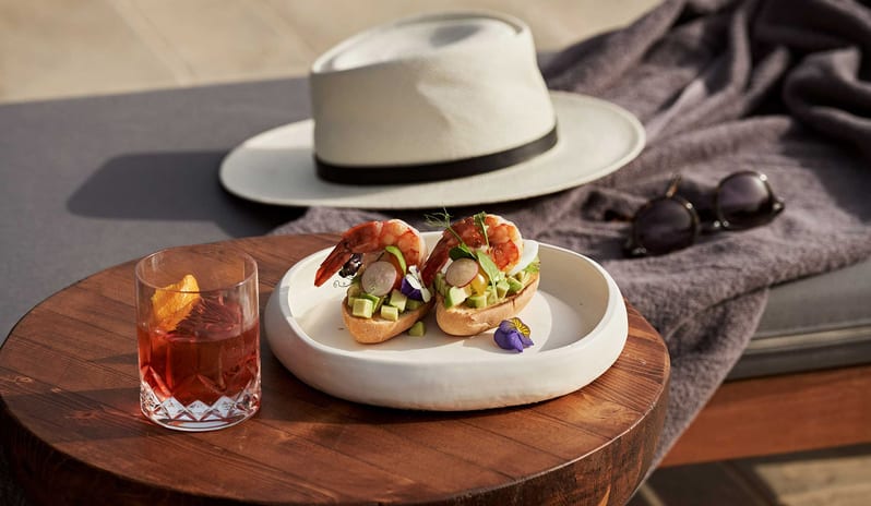 The Spiritzeria tGourmet shrimp and avocado appetizers on a wooden table next to a Negroni cocktail, featuring a white sun hat and sunglasses at Aulūs Lindos Rhodes, Curio Collection by Hilton by Kenwood Travel