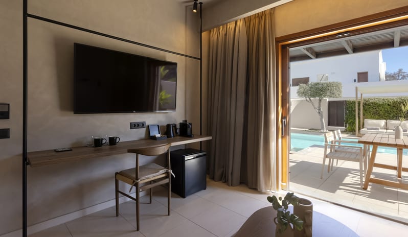 Tropical Bungalow Suite with Private Pool a wall-mounted TV over a wooden desk, a mini-fridge, and large sliding doors opening to a private pool and outdoor dining areaat Aulūs Lindos Rhodes, Curio Collection by Hilton by Kenwood Travel