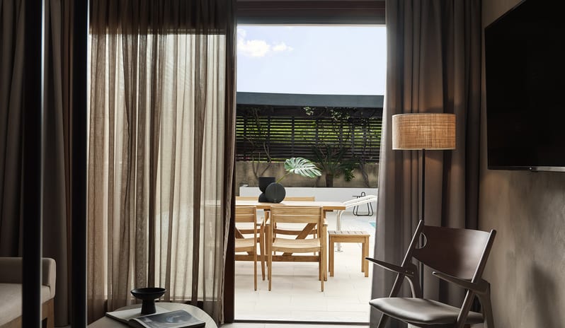 Tropical Pavilion One Bedroom Suite with Private Pool wooden slatted lounge chair, linen curtains, and glass doors opening to a lush private garden at Aulūs Lindos Rhodes, Curio Collection by Hilton by Kenwood Travel