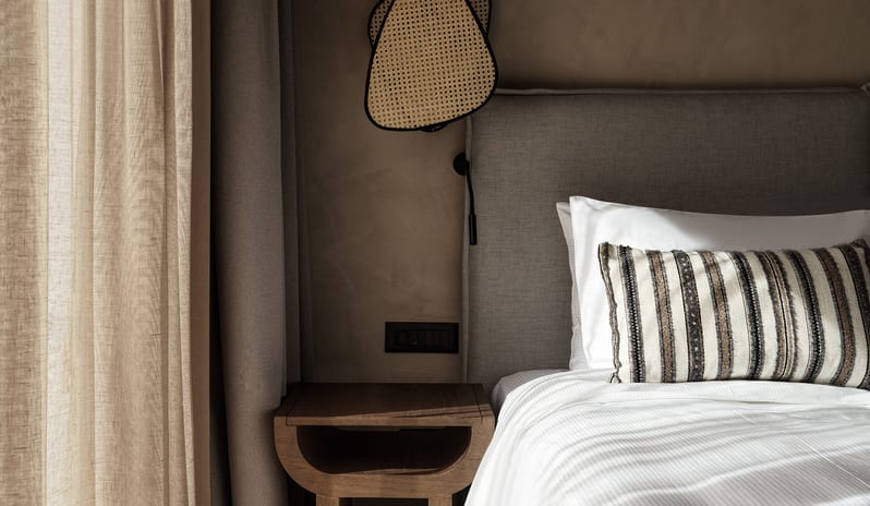 Tropical Pavilion Two Bedroom Suite with Outdoor Jacuzzi luxury bed with a grey upholstered headboard, a striped accent pillow, and a unique woven rattan light fixture next to a wooden bedside table at Aulūs Lindos Rhodes, Curio Collection by Hilton by Kenwood Travel