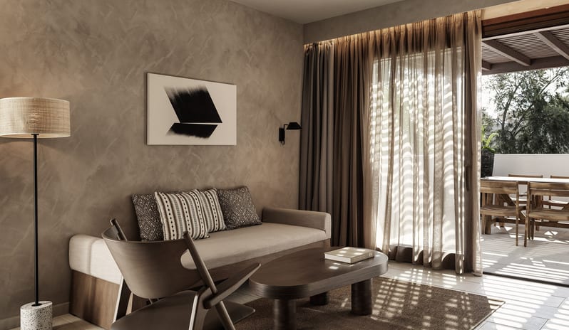 Tropical Pavilion Two Bedroom Suite with Outdoor Jacuzzi living room with a neutral-toned sofa, dark wood coffee table, and textured walls, leading to an outdoor dining area on a shaded patio at Aulūs Lindos Rhodes, Curio Collection by Hilton by Kenwood Travel