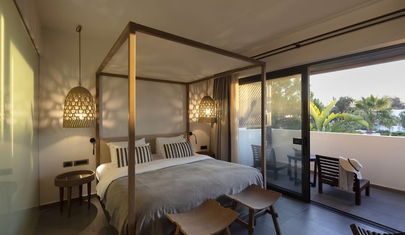 Tropical Zen Bungalow Suite Bedroom With a Canopy Bed, Stylish Rattan Lamps, and Stone Tile Flooring at Aulūs Lindos Rhodes, Curio Collection by Hilton by Kenwood Travel