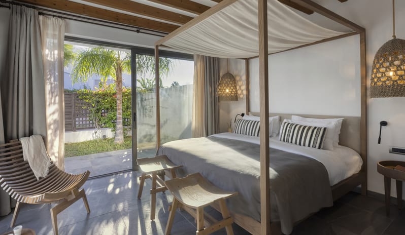 Tropical Zen Bungalow Suite with Private Pool bedroom with a wooden canopy bed, slatted lounge chair, and sliding glass doors opening to a private garden at Aulūs Lindos Rhodes, Curio Collection by Hilton by Kenwood Travel