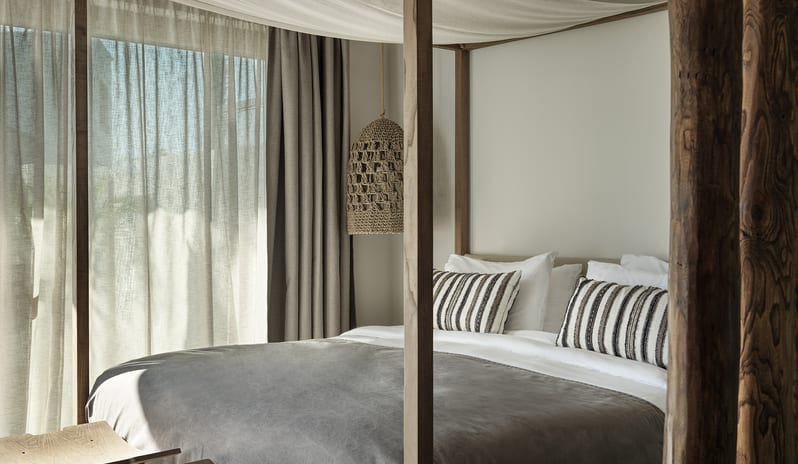 Tropical Zen Bungalow Suite with Private Pool bedroom with a wooden canopy bed, slatted lounge chair, and sliding glass doors opening to a private garden at Aulūs Lindos Rhodes, Curio Collection by Hilton by Kenwood Travel