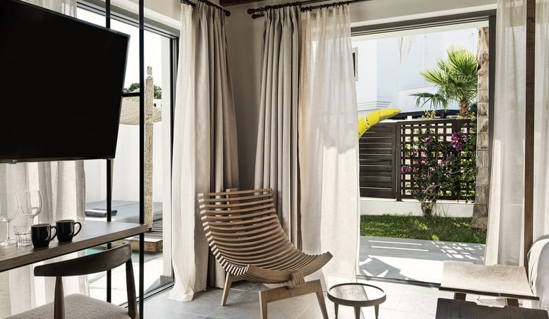 Tropical Zen Bungalow Suite with Private Pool wooden slatted lounge chair, canopy bed, and floor-to-ceiling glass doors opening to a private patio with palm trees. at Aulūs Lindos Rhodes, Curio Collection by Hilton by Kenwood Travel