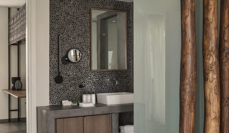 Tropical Zen Bungalow Suite with Private Pool bathroom featuring a dark pebble mosaic accent wall, a gray concrete vanity with a white vessel sink, and rustic wood log room dividers at Aulūs Lindos Rhodes, Curio Collection by Hilton by Kenwood Travel