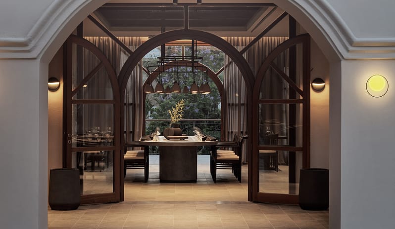 Yellow Sea Restaurant (Adults Only) entrance featuring a white stucco archway leading into a warm, lit dining room with a large communal table at Aulūs Lindos Rhodes, Curio Collection by Hilton by Kenwood Travel