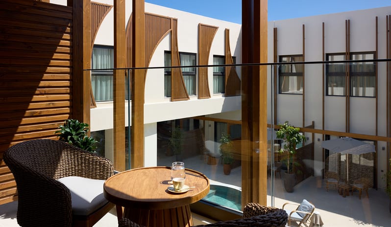 Atrium Escape tropical palm trees, rattan lounge chairs, and a small table with cocktails overlooking a modern white building at Aulūs Chania, Curio Collection by Hilton by Kenwood Travel