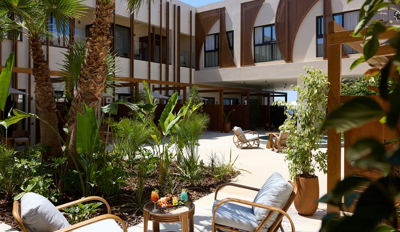 Atrium Escape tropical palm trees, rattan lounge chairs, and a small table with cocktails overlooking a modern white building at Aulūs Chania, Curio Collection by Hilton by Kenwood Travel