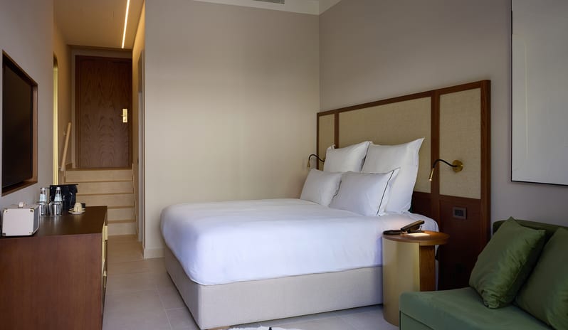 Atrium Escape Bedroom featuring a white plush bed, wood-paneled headboard, and a green velvet armchair at Aulūs Chania, Curio Collection by Hilton by Kenwood Travel