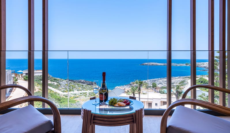Sea View Escape glass-railed balcony with two wooden armchairs and a small table set with champagne and fruit, overlooking a rocky blue coastline at Aulūs Chania, Curio Collection by Hilton by Kenwood Travel