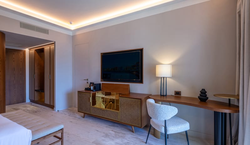 Tropical Escape bedside, showing a large sliding glass door that opens to a private balcony with two wicker lounge chairs at Aulūs Chania, Curio Collection by Hilton by Kenwood Travel