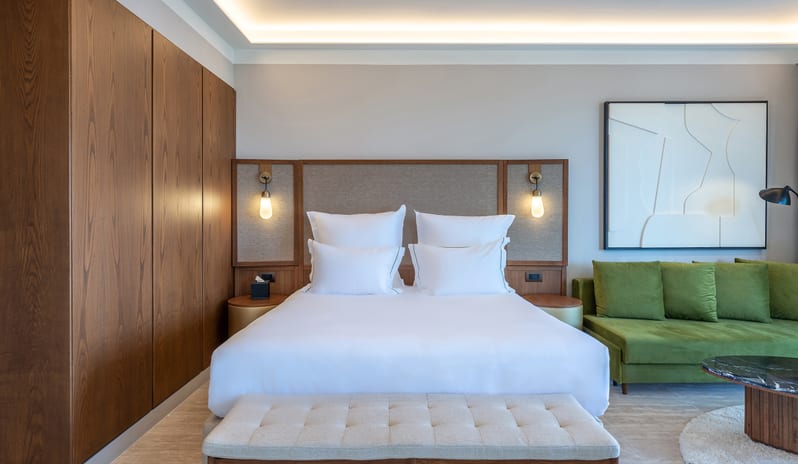 Tropical Escape Bedroom featuring a king-sized bed with white linens, a walnut wood paneled wall, and a moss green velvet sofa at Aulūs Chania, Curio Collection by Hilton by Kenwood Travel