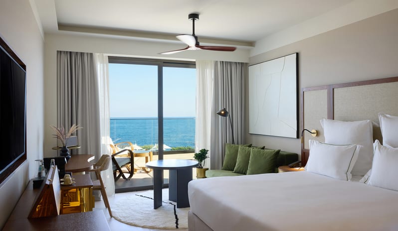 Infinite Sea View Escape Bedroom featuring a white king-size bed, sage green velvet sofa, and a private balcony overlooking a blue ocean at Aulūs Chania, Curio Collection by Hilton by Kenwood Travel