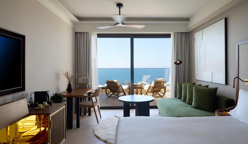 Infinite Sea View Escape Bedroom featuring a white king-size bed, sage green velvet sofa, and a private balcony overlooking a blue ocean at Aulūs Chania, Curio Collection by Hilton by Kenwood Travel
