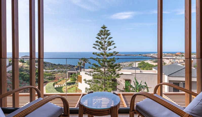 Sea View Escape glass-railed balcony with two wooden armchairs and a small table set with champagne and fruit, overlooking a rocky blue coastline at Aulūs Chania, Curio Collection by Hilton by Kenwood Travel