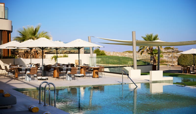 Tesoro Mio Restaurant poolside restaurant featuring white sun umbrellas, wicker dining sets, and shade sails under a bright blue sky at Aulūs Chania, Curio Collection by Hilton by Kenwood Travel