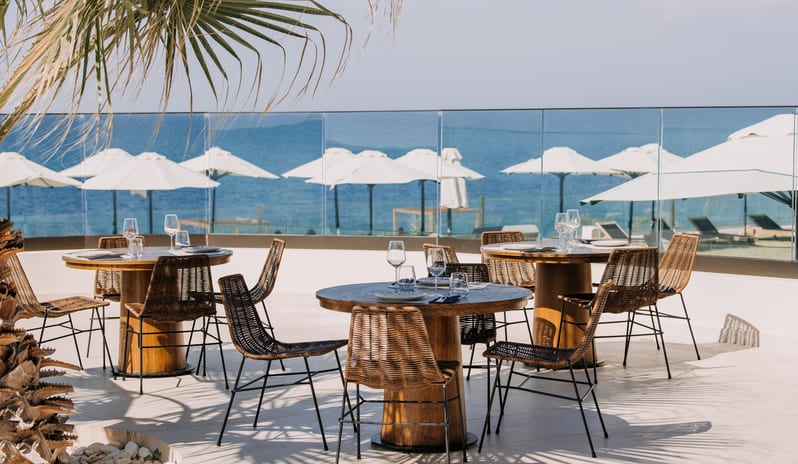 Tesoro Mio Restaurant Sunlit outdoor dining deck overlooking the sea, featuring round wooden tables with woven wicker chairs and white beach umbrellas at Aulūs Chania, Curio Collection by Hilton by Kenwood Travel