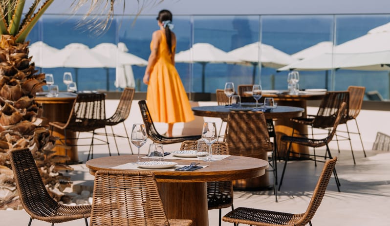 Tesoro Mio Restaurant Sunlit outdoor dining deck overlooking the sea, featuring round wooden tables with woven wicker chairs and white beach umbrellas at Aulūs Chania, Curio Collection by Hilton by Kenwood Travel