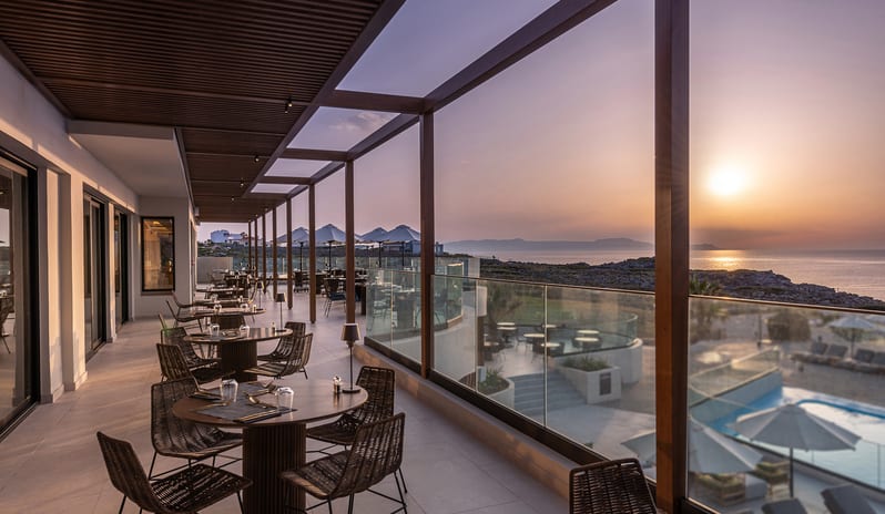 The Island Buffet Restaurant terrace at sunset featuring modern woven chairs, round wooden tables, and a glass railing overlooking the ocean and a resort swimming pool at Aulūs Chania, Curio Collection by Hilton by Kenwood Travel