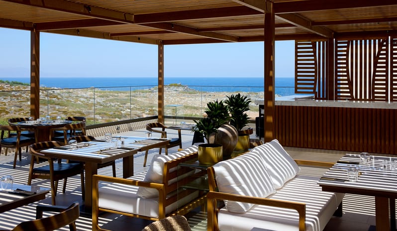 The Island Buffet Restaurant terrace under a wooden pergola with striped shadows falling across white outdoor sofas, wooden dining sets at Aulūs Chania, Curio Collection by Hilton by Kenwood Travel
