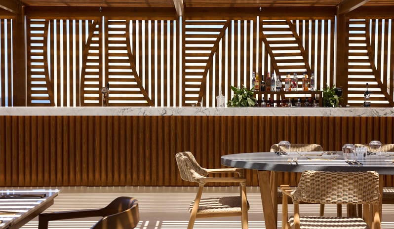 The Island Buffet Restaurant terrace under a wooden pergola with striped shadows falling across white outdoor sofas, wooden dining sets at Aulūs Chania, Curio Collection by Hilton by Kenwood Travel