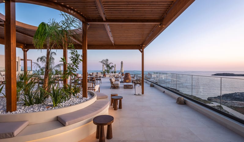 The Spiritzeria bar and lounge featuring a wooden pergola, circular stone planters with greenery, and elegant seating overlooking a calm ocean at sunset at Aulūs Chania, Curio Collection by Hilton by Kenwood Travel