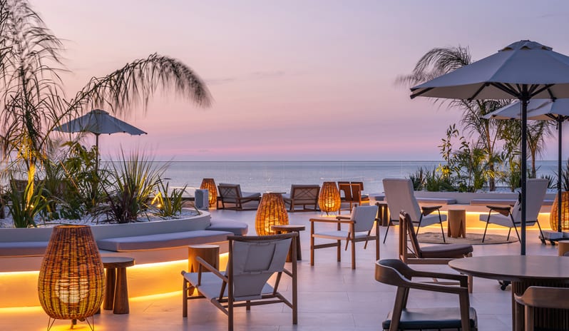 The Spiritzeria bar and lounge featuring a wooden pergola, circular stone planters with greenery, and elegant seating overlooking a calm ocean at sunset at Aulūs Chania, Curio Collection by Hilton by Kenwood Travel