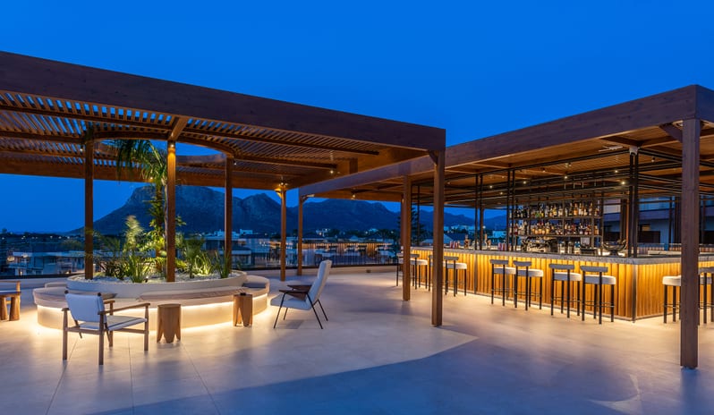 The Spiritzeria bar and lounge featuring a wooden pergola, circular stone planters with greenery, and elegant seating overlooking a calm ocean at sunset at Aulūs Chania, Curio Collection by Hilton by Kenwood Travel
