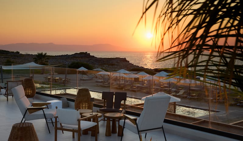 The Spiritzeria bar and lounge featuring a wooden pergola, circular stone planters with greenery, and elegant seating overlooking a calm ocean at sunset at Aulūs Chania, Curio Collection by Hilton by Kenwood Travel