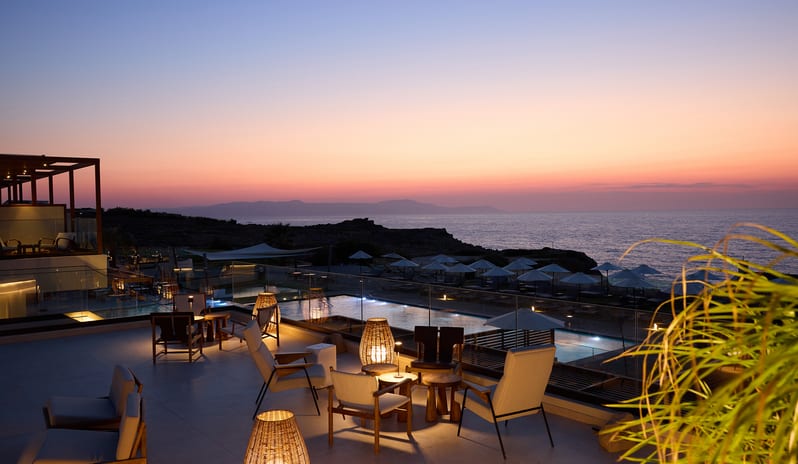 The Spiritzeria bar and lounge featuring a wooden pergola, circular stone planters with greenery, and elegant seating overlooking a calm ocean at sunset at Aulūs Chania, Curio Collection by Hilton by Kenwood Travel