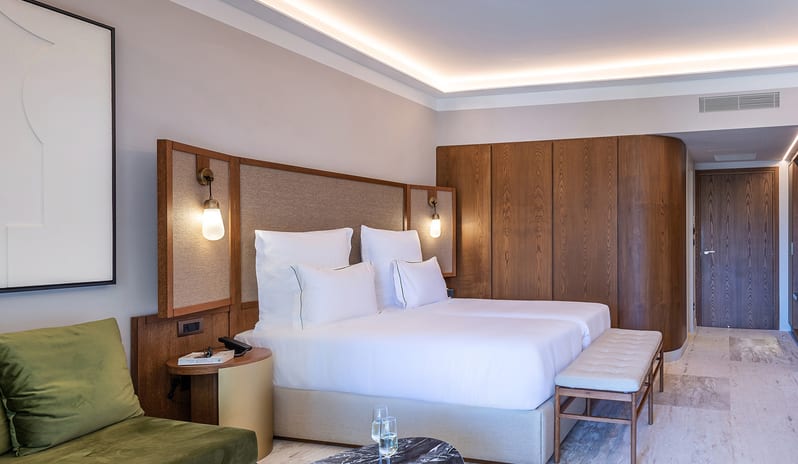 Tropical Open Plan Suite (with shared pool) Bedroom with white linens, a curved wood-paneled wall, green velvet seating, and marble accents at Aulūs Chania, Curio Collection by Hilton by Kenwood Travel