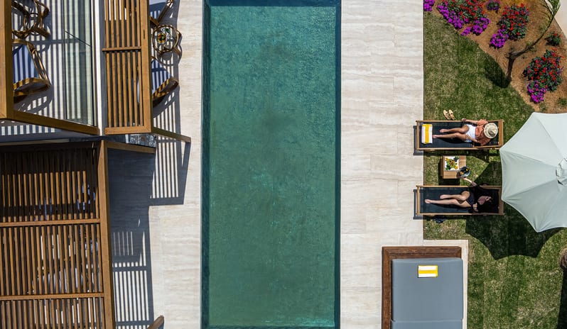 Tropical Open Plan Suite (with shared pool) swimming pool flanked by a wooden pergola, a stone patio, and two people relaxing on lounge chairs at Aulūs Chania, Curio Collection by Hilton by Kenwood Travel