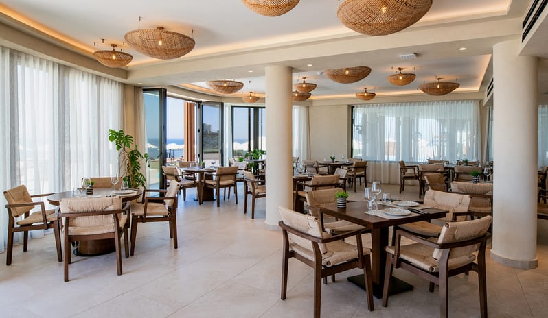 Yellow Sea Restaurant interior featuring woven rattan pendant lights, wooden dining tables with woven chairs and large folding glass doors opening to a seaside view at Aulūs Chania, Curio Collection by Hilton by Kenwood Travel