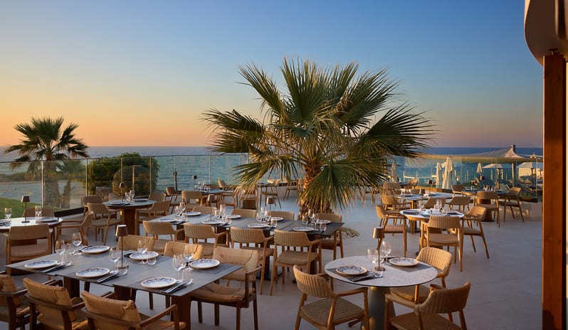 Yellow Sea Restaurant dining area featuring a burnt-orange terracotta ribbed booth, dark wood tables set with patterned plates and wine glasses at Aulūs Chania, Curio Collection by Hilton by Kenwood Travel