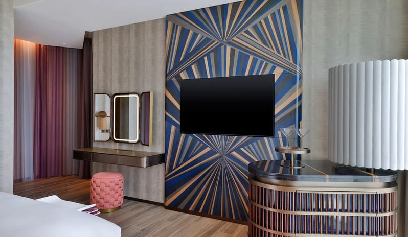 Room interior featuring a wall-mounted TV on a blue geometric patterned backdrop, a vanity desk with a lighted mirror and pink ottoman, and a stylish minibar unit at W Dubai - Mina Seyahi by Kenwood Travel