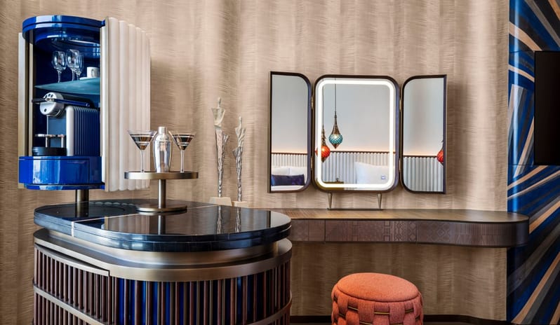 Room interior featuring a wall-mounted TV on a blue geometric patterned backdrop, a vanity desk with a lighted mirror and pink ottoman, and a stylish minibar unit at W Dubai - Mina Seyahi by Kenwood Travel