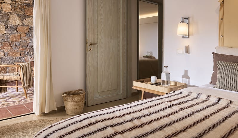 Three Bedroom Thalassa Villa Private Pool Seafront bed with a striped throw, a wooden tray with refreshments, and an open door leading to a rustic stone-walled terrace at St.Nicolas Bay Resort by Kenwood Travel