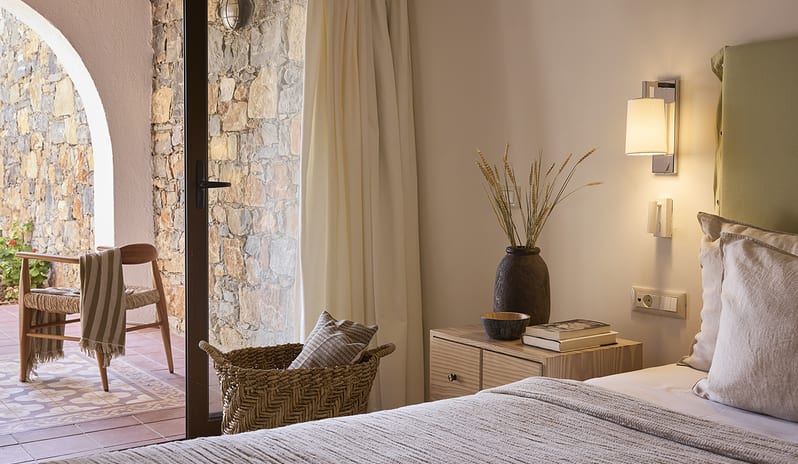 Three Bedroom Thalassa Villa Private Pool Seafront wooden bedside table with books, a woven basket, and an open doorway leading to a stone-walled private terrace at St.Nicolas Bay Resort by Kenwood Travel