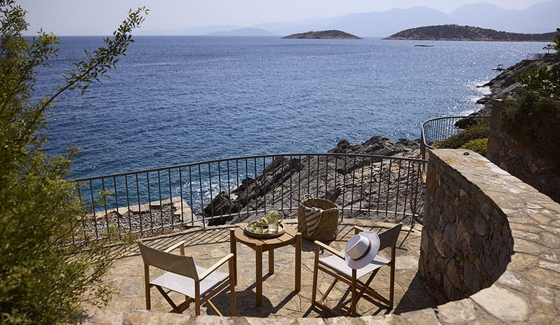 Three Bedroom Thalassa Villa Private Pool Seafront wooden bistro table, directors' chairs, and a white sun hat, overlooking a rocky coastline and the Mediterranean Sea at St.Nicolas Bay Resort by Kenwood Travel