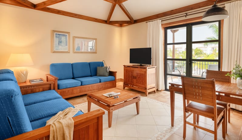 Suite living room featuring blue sofas, vaulted wooden ceiling beams, and a dining area with a balcony view at Princesa Yaiza Suite Hotel Resort by Kenwood Travel