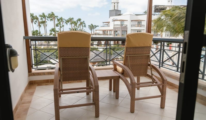 Suite Relax balcony with two wicker lounge chairs and a small table, overlooking a resort courtyard with palm trees and white buildings at Princesa Yaiza Suite Hotel Resort by Kenwood Travel