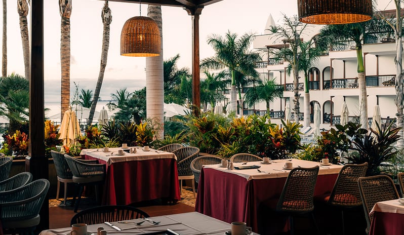 Outdoor dining terrace at Hacienda restaurant with red tablecloths, wicker lamps, and palm tree views at Princesa Yaiza Suite Hotel Resort in Europe Holidays by Kenwood Travel