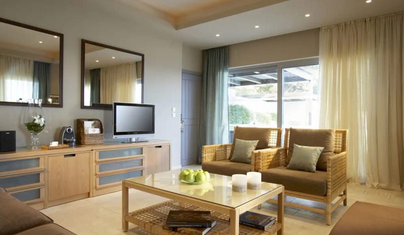 Presidential Bungalow woven armchairs, a glass-top coffee table with green apples, a flat-screen TV, and large mirrors reflecting soft neutral curtains at Eagles Palace by Kenwood Travel