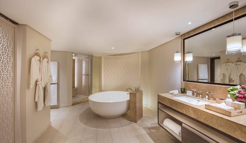Palm Suite bathroom featuring a large white freestanding soaking tub, a double marble vanity with modern fixtures, hanging robes, and a walk-in glass shower under soft, warm lighting at Royal Palm Beachcomber Luxury by Kenwood Travel