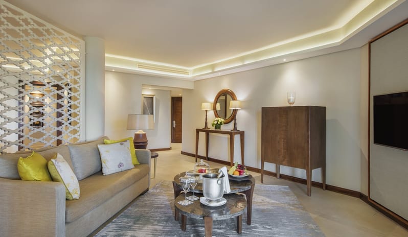 Palm Suite living room featuring a plush sofa, circular marble coffee tables with champagne and fruit, a wall-mounted TV, and elegant dark wood furniture accents at Royal Palm Beachcomber Luxury by Kenwood Travel