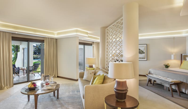 Palm Suite living area with a plush sofa, marble coffee table, and fruit platter, separated by a decorative white screen from a bedroom area at Royal Palm Beachcomber Luxury by Kenwood Travel