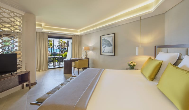 Palm Suite king-sized bed with lime green accent pillows, a dedicated workspace, and large sliding glass doors opening onto a private terrace with a view of tropical palms and the ocean at Royal Palm Beachcomber Luxury by Kenwood Travel
